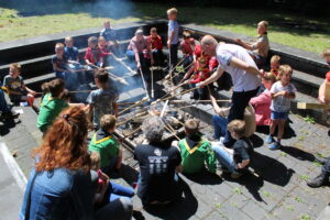 Foto van een kampvuur waar kinderen omheen zitten om een broodje op een lange stok te bakken.